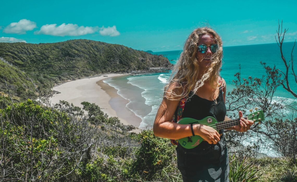 Smiling woman playing a green ukulele on a cliffside overlooking a tropical beach and ocean under a clear blue sky.
