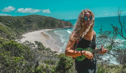 Smiling woman playing a green ukulele on a cliffside overlooking a tropical beach and ocean under a clear blue sky.