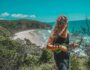 Smiling woman playing a green ukulele on a cliffside overlooking a tropical beach and ocean under a clear blue sky.