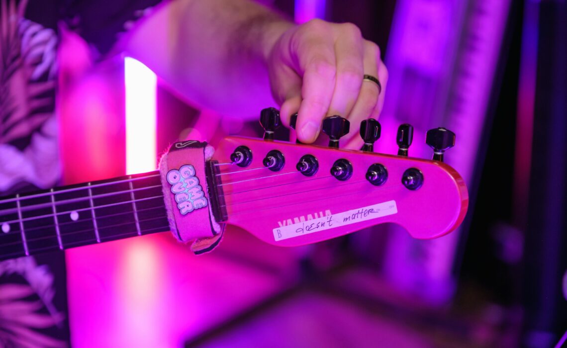 A person tuning a red Yamaha electric guitar under vibrant purple stage lights, with a "Game Over" fret wrap on the neck.