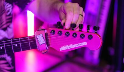 A person tuning a red Yamaha electric guitar under vibrant purple stage lights, with a "Game Over" fret wrap on the neck.