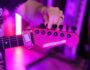A person tuning a red Yamaha electric guitar under vibrant purple stage lights, with a "Game Over" fret wrap on the neck.