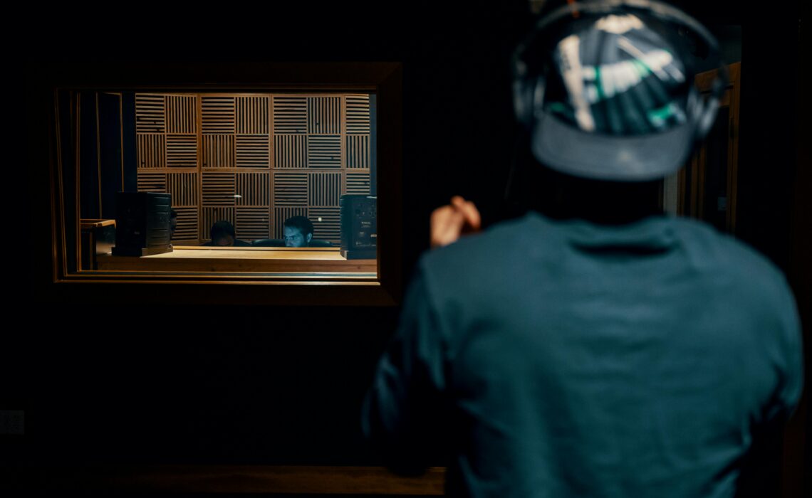 A person wearing headphones stands in a recording studio, facing a soundproof window with two audio engineers working behind a mixing desk.