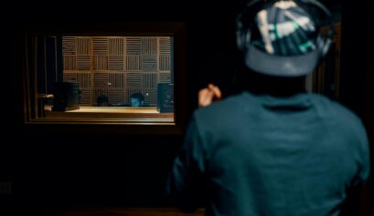 A person wearing headphones stands in a recording studio, facing a soundproof window with two audio engineers working behind a mixing desk.