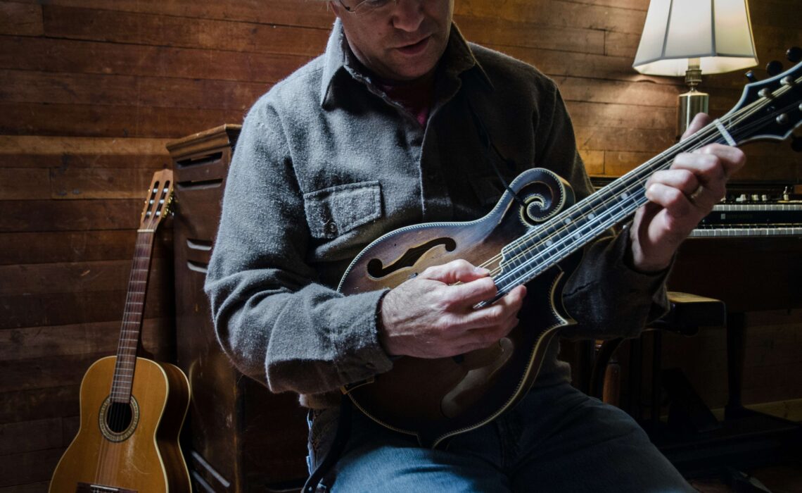 Musician playing a mandolin in a cozy wooden room with a classical guitar resting nearby.