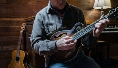 Musician playing a mandolin in a cozy wooden room with a classical guitar resting nearby.