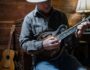 Musician playing a mandolin in a cozy wooden room with a classical guitar resting nearby.