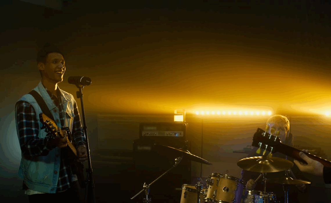 A band performing on a small stage with warm yellow backlighting, featuring a smiling guitarist singing into a microphone, a drummer in the background, and dramatic stage lighting creating depth