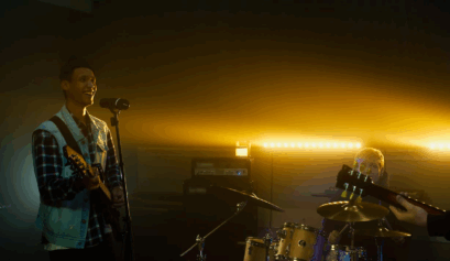 A band performing on a small stage with warm yellow backlighting, featuring a smiling guitarist singing into a microphone, a drummer in the background, and dramatic stage lighting creating depth