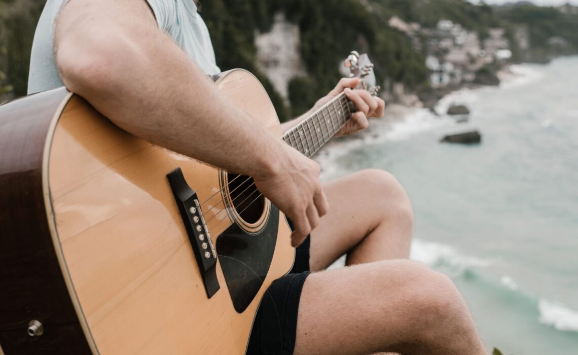 Person playing an acoustic guitar overlooking the ocean, practicing easy summer songs on guitar by the beach.