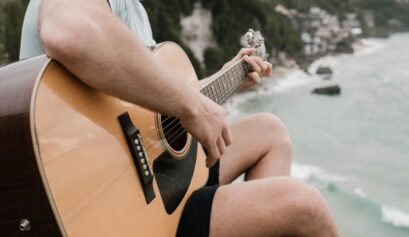 Person playing an acoustic guitar overlooking the ocean, practicing easy summer songs on guitar by the beach.