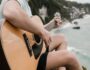 Person playing an acoustic guitar overlooking the ocean, practicing easy summer songs on guitar by the beach.
