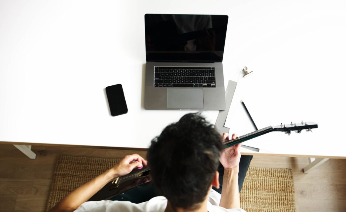 Guitarist practicing acoustic guitar at home beside a laptop, surrounded by everyday music and practice essentials.