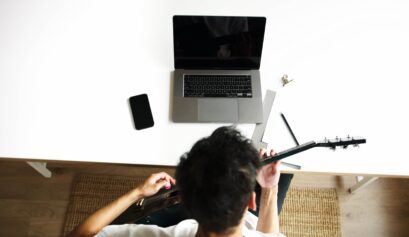 Guitarist practicing acoustic guitar at home beside a laptop, surrounded by everyday music and practice essentials.